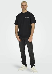 T-shirt nera in cotone con maniche corte e un piccolo logo bianco sul lato sinistro del petto, abbinata a jeans scuri e sneakers bianche.