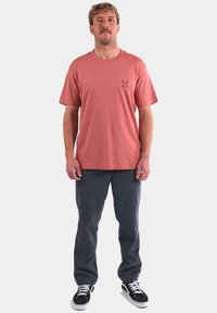 Camiseta de algodón rosa con un pequeño gráfico en el lado izquierdo del pecho, combinada con pantalones gris oscuro y zapatillas negras con suelas blancas.