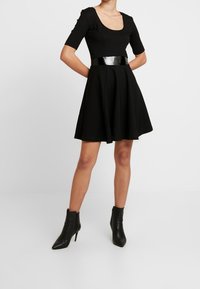 Svart klänning med korta ärmar, båtformat halsringning och ett glansigt svart bälte. Utsågd kjoldesign ihop med spetsiga svarta ankelboots.