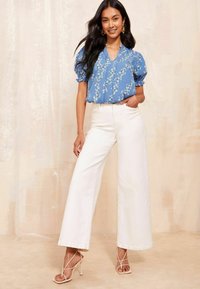 Donna che indossa una blusa corta a maniche corte blu a fiori infilata in pantaloni bianchi a gamba larga a vita alta e sandali con tacco a strappy beige, sorridendo.
