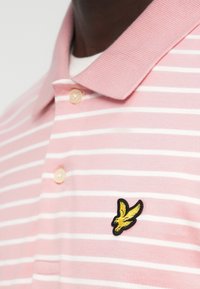 Ροζ ριγέ πόλο μπλούζα με ριμπ ύφασμα στον γιακά, με κίτρινο κεντητό λογότυπο πουλιού στο στήθος και κουμπιά από πέρλα.