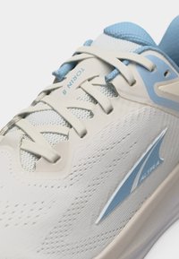 Κοντινό πλάνο άσπρου αθλητικού παπουτσιού με ανοιχτά μπλε λεπτομέρειες, μπεζ κορδόνια και ετικέτα "TORIN 8" στη γλώσσα.