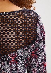 Overlay in pizzo nero con un motivo floreale in rosso e bianco. Retro trasparente con un design geometrico e bordo curvato. Tessuto morbido e texturizzato.