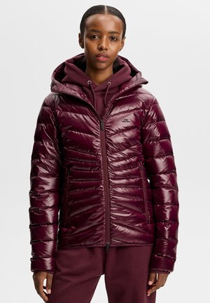 Vrouw draagt een glanzende bordeauxrode pufferjas en bijpassende hoodie, staand met een ontspannen houding tegen een effen achtergrond.