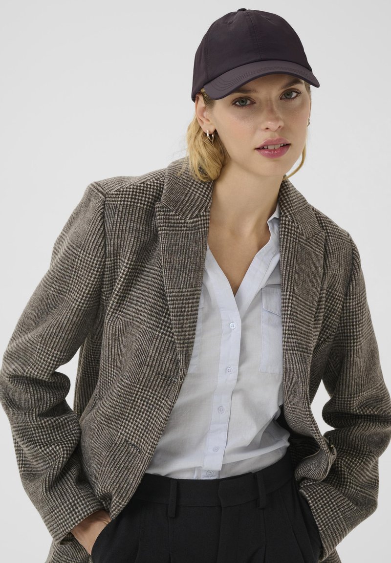 Bruine geblokte blazer met een gestructureerd ontwerp, een lichtblauwe overhemden en een zwarte baseballcap. Getextureerde stof en pasvorm op maat.