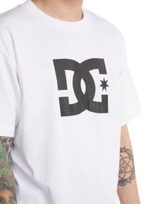 Weißes Baumwoll-T-Shirt mit großem schwarzem DC-Logo und Stern-Design. Kurzarm und Rundhalsausschnitt mit glatter Textur.