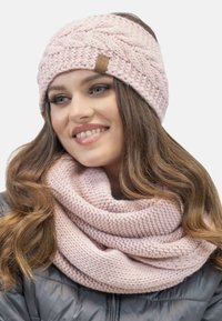 Rosa stickad mössa med flätat mönster och en läderetikett, kombinerad med en matchande texturerad halsduk, bärs över en mörk vadderad jacka.