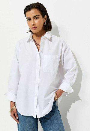 Jonge vrouw met kort donker haar, dragend een losse witte blouse met knopen en een blauwe spijkerbroek, staand met één hand in de zak tegen een witte achtergrond.