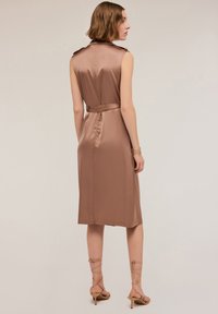 Rochie din satin maro cu design fără mâneci, talie accentuată cu curea și tiv până la genunchi. Prezintă epoleți la umeri și o textură netedă. Poartă pantofi cu barete.