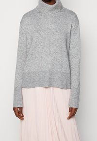 Donkerhuidige persoon in een lichtgrijze gebokte coltrui en een lichtroze geplooide rok, afgebeeld van de schouders tot halverwege de dijen.