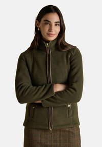 Žena s dlhými tmavými vlasmi, na sebe má zlaté kruhové náušnice, olivovozelenú fleecovú bundu so zipsom a hnedú kockovanú sukňu, so prekríženými rukami.