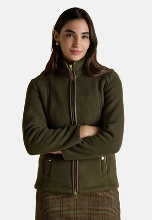Frau mit langen, dunklen Haaren, die goldene Creolen trägt, eine olivgrüne Reißverschluss-Fleecejacke und einen braun karierten Rock, die Arme verschränkt.