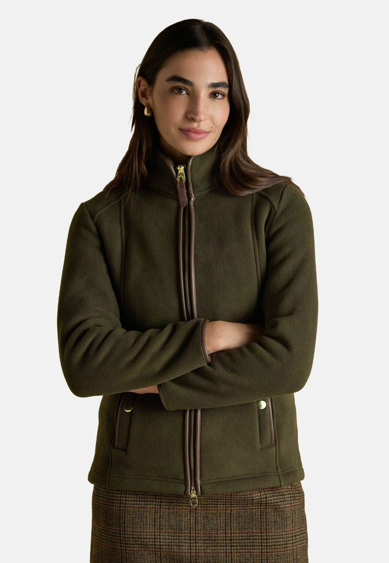 Žena s dlhými tmavými vlasmi, na sebe má zlaté kruhové náušnice, olivovozelenú fleecovú bundu so zipsom a hnedú kockovanú sukňu, so prekríženými rukami.