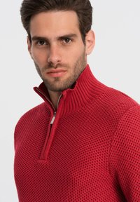 Rode gebreide ritsvest met een hoge kraag, voorzien van een gestructureerd patroon en een zilveren rits. De aansluitende pasvorm verhoogt de warmte.