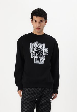 Sweatshirt noir avec un graphisme en couches blanc et gris "KARL LAGERFELD", col rond et poignets côtelés. Associé à un pantalon noir à motifs.