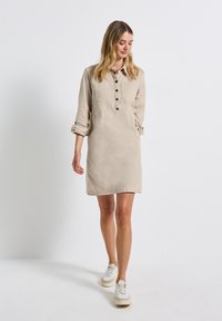 Robe chemise beige clair avec un col, une fermeture à boutons sur le devant et deux poches poitrine ; conçue dans un style décontracté avec des manches retroussées.