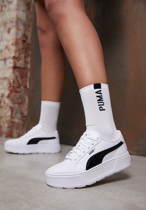 Baskets à plateforme blanches avec accents noirs, dotées d'une semelle texturée et d'un design à lacets. Portées avec des chaussettes blanches montantes affichant un logo.