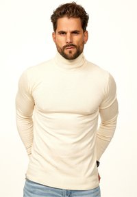 Cremfarbener Rollkragenpullover mit langen Ärmeln, taillierter Passform und geripptem Saum, getragen von einem bärtigen Mann. Schlichte Textur, keine Muster.