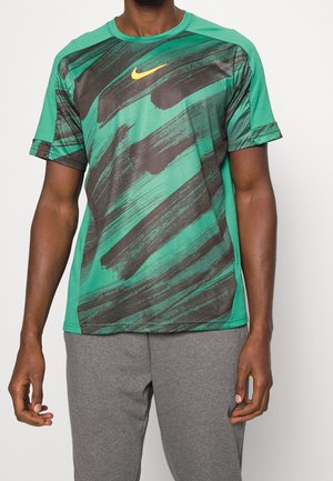 Mann trägt grünes und schwarzes Nike-Sportshirt mit abstraktem Muster und graue Nike-Sweathose, steht vor einfarbigem Hintergrund.