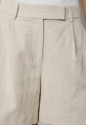 Prim-plan cu pantaloni scurți din țesătură bej cu model chevron, având bucle pentru curea și pliseuri frontale, purtați cu un top alb.