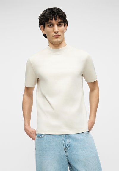 Άνδρας με σγουρά σκούρα μαλλιά, που φοράει κοντομάνικο μπεζ πουκάμισο με ψηλό γιακά και ανοιχτό μπλε τζιν, με τα χέρια στις τσέπες, στέκεται μπροστά σε απλό φόντο.