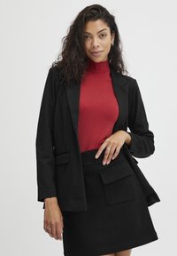 Černý blazer s klopami, spárovaný s červeným rolákem a černou sukní s přední kapsou. Texturovaná tkanina, elegantní design.