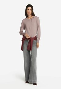 Helles rosa langärmliges Oberteil, kombiniert mit grauen Jeans mit weitem Bein. Ein bordeauxroter Pullover ist um die Taille gebunden. Glattes Material, minimalistisches Design.