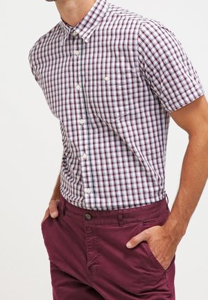 Hombre vistiendo una camisa blanca de manga corta con estampado a cuadros rojo y negro, combinada con pantalones burdeos, con las manos en los bolsillos.