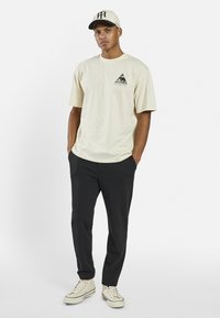 Beige cotton t-shirt with a triangular eye graphic on the left chest, paired with black pants and cream sneakers. Cap features a logo on the front.