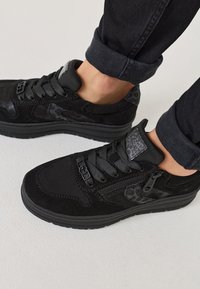 Fekete sneaker, amely velúr és texturált anyagok keverékéből készült, sima dizájnnal, fűzős rögzítéssel és oldalsó cipzár részlettel.