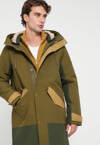 Parka vert kaki avec capuche doublée de fourrure, fermeture éclair à l'avant, poches doubles et accents beiges sur les poignets et les boutons.