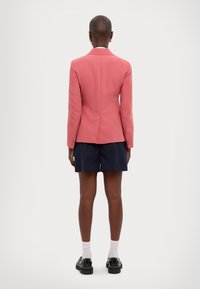 Blazer rosa su misura con spalle strutturate, spacco centrale e polsini con bottoni, abbinato a pantaloni navy e scarpe nere.