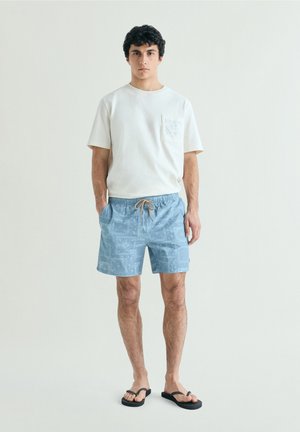 Joven con camisa blanca de manga corta, pantalones cortos estampados de color azul claro con cordón y chanclas negras, de pie contra un fondo liso.