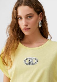 Jeune femme aux cheveux bruns ondulés portant une chemise jaune pâle avec une broche ornée de bijoux et de grands boucles d'oreilles argentées réfléchissantes sur un fond blanc.