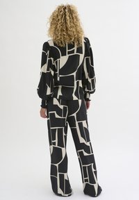 Person with curly blonde hair wearing matching black and beige geometric patterned blouse with puff sleeves and wide-leg pants, standing facing away.