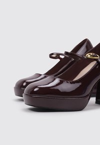 Zapatos Mary Jane de charol burdeos con punta redonda, correa en el tobillo y tacón grueso. Textura suave con un acabado brillante.