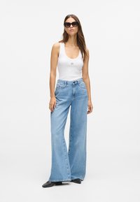 Ανοιχτό μπλε φαρδύ τζιν με ψηλή μέση, συνδυασμένο με λευκό μπλουζάκι και μαύρα γυαλιά ηλίου, με ασημένια τρουκς στα τζιν.