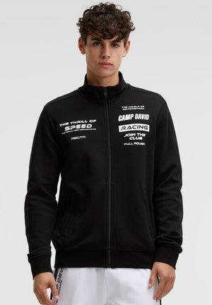 Young man with curly hair wearing a black zip-up jacket with white racing-themed text and white pants against a plain background.