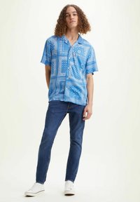Camisa de manga corta azul con estampado blanco; combinada con unos vaqueros ajustados azules y zapatillas blancas; estilo casual, corte relajado.