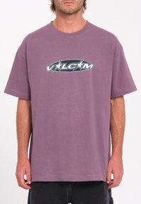 T-shirt en coton violet à manches courtes avec un design de logo graphique en noir et blanc sur le devant. Coupe décontractée, col rond.