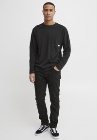 Black long-sleeve T-shirt with a chest pocket paired with black slim-fit pants and black sneakers featuring white stripes and soles.