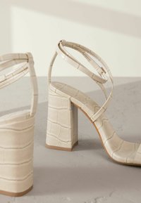 Sandales à talons hauts beige avec un motif crocodile texturé, talon bloc rectangulaire et fines lanières croisées autour de la cheville.