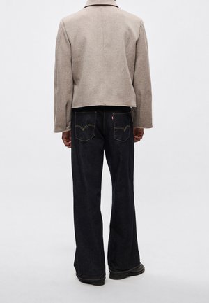 Personne portant une veste courte beige et un jean large foncé, debout dos à la caméra sur un fond uni.