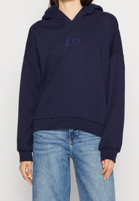Sudadera azul marino hecha de tejido suave, con un bolsillo frontal, ajuste oversize y logo de GAP bordado en el pecho. Combinada con jeans de un azul claro.