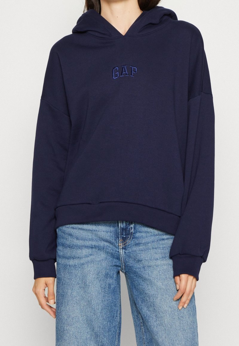 Sudadera azul marino hecha de tejido suave, con un bolsillo frontal, ajuste oversize y logo de GAP bordado en el pecho. Combinada con jeans de un azul claro.