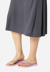 Piedi che indossano infradito intrecciate di colore viola chiaro con cinturini luccicanti, appoggiati su uno sfondo neutro, con una gonna grigia scura visibile sopra.