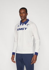 Baltas ilgomis rankovėmis polo marškinėlis mėlynu apykaklės ir rankogalio akcentu. Turi vertikalius ribinius raštus ir krūtinėje mėlynu užrašytą „ΕΜΣΤ“.