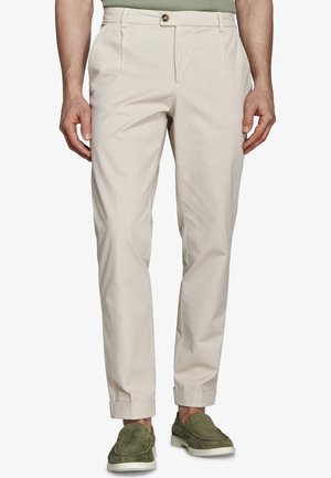 Mężczyzna w beżowych plisowanych spodniach z mankietami i zielonych zamszowych butach wsuwanych, stojący na białym tle.