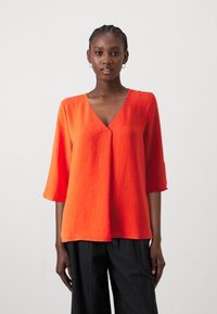 Blusa rossa, con texture, a scollatura a V, maniche tre quarti e vestibilità rilassata, abbinata a pantaloni neri e larghi.