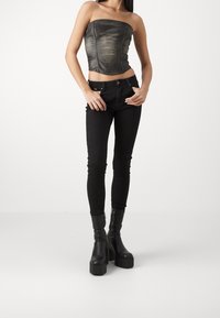 Top senza spalline in pelle nera con cuciture curve, abbinato a jeans skinny neri e stivali neri con tacco grosso, in piedi davanti a uno sfondo bianco.